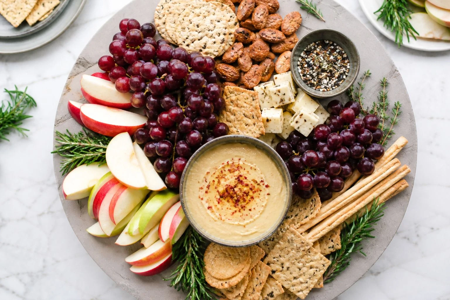 High-protein snack platter with hummus, grapes, apple slices, crackers, nuts, cheese, and rosemary.