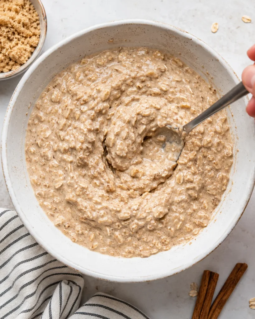 Creamy brown sugar overnight oat mixture being stirred in a bowl with cinnamon flecks and hydrated oats