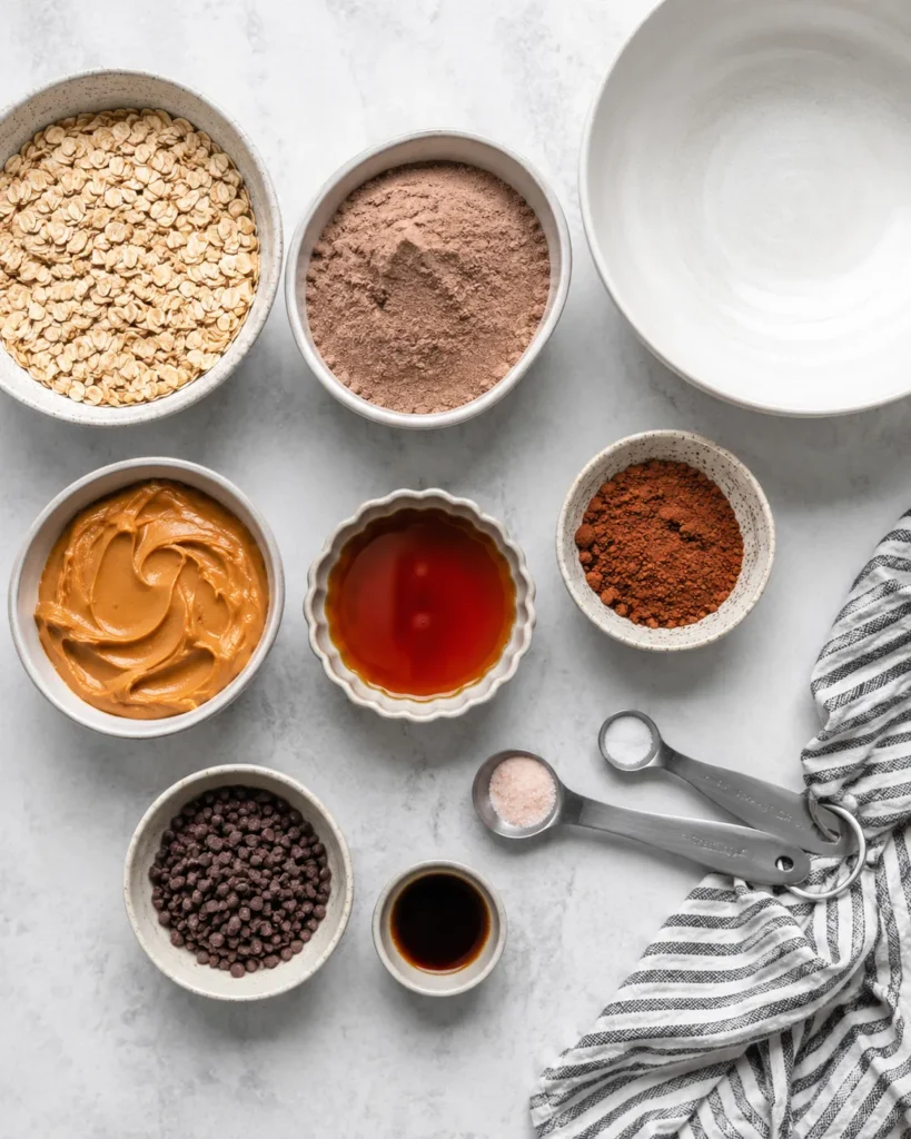 Ingredients arranged for making brownie protein bites with oats, cocoa, protein powder, peanut butter, maple syrup, and chocolate chips
