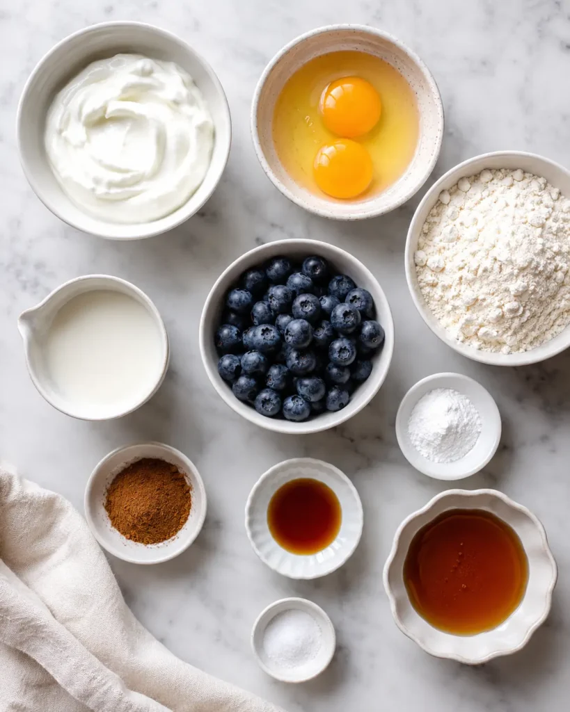 Ingredients for Greek yogurt protein pancakes, including yogurt, eggs, flour, milk, blueberries, maple syrup, and spices