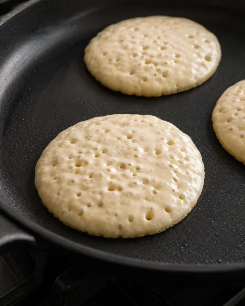 Protein pancakes cooking in a skillet with bubbles on top and edges beginning to set